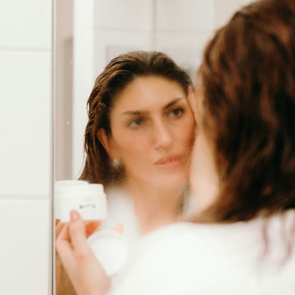 A woman in a bathroom holds Radford Butter -  The Thick Cream jar while looking into a mirror, with her reflection showing wet hair and a soft-focus view of the room.
