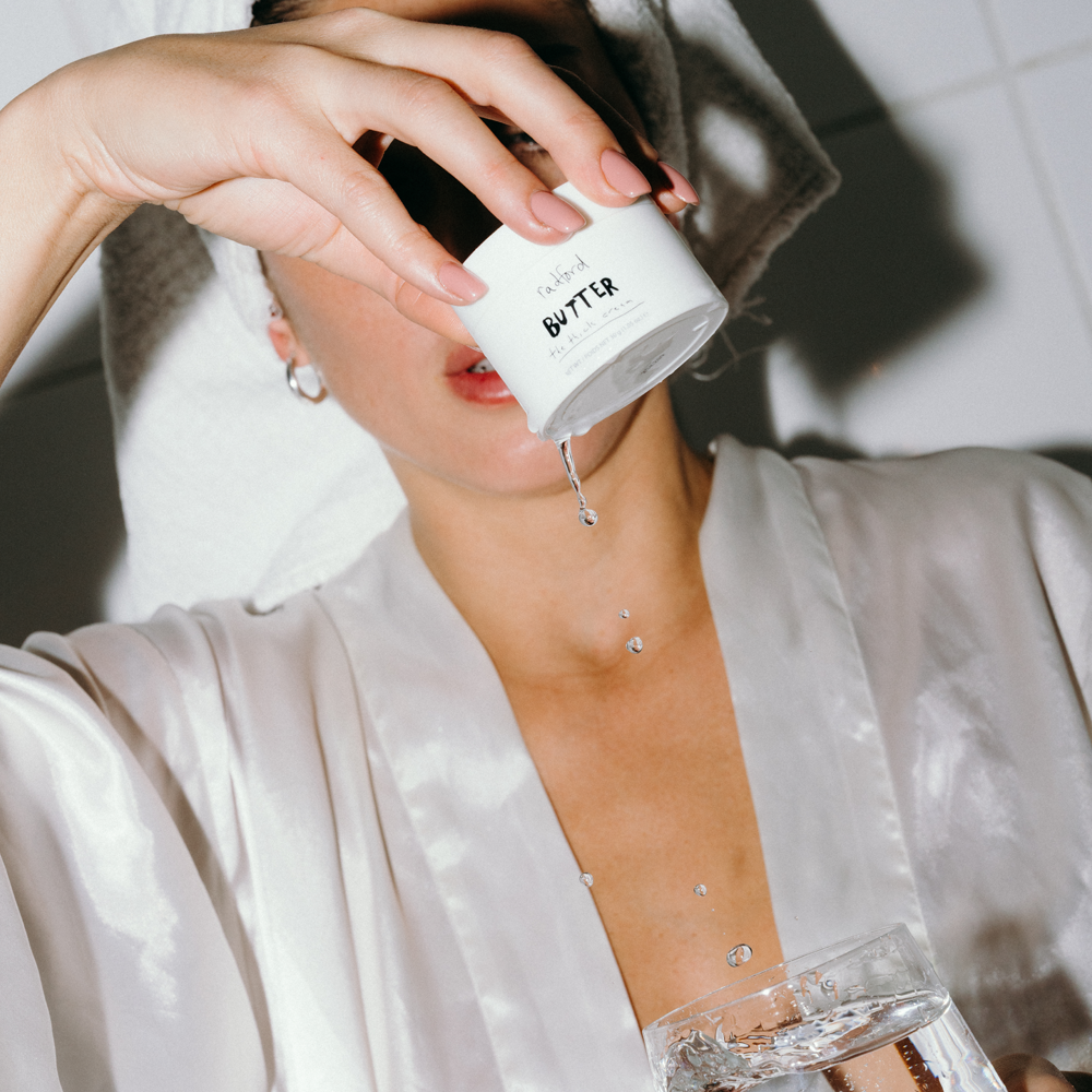 A woman in a white satin robe holds a white jar of Radford Butter -  The Thick Cream above her face, with product dripping water into a glass of water in the foreground.