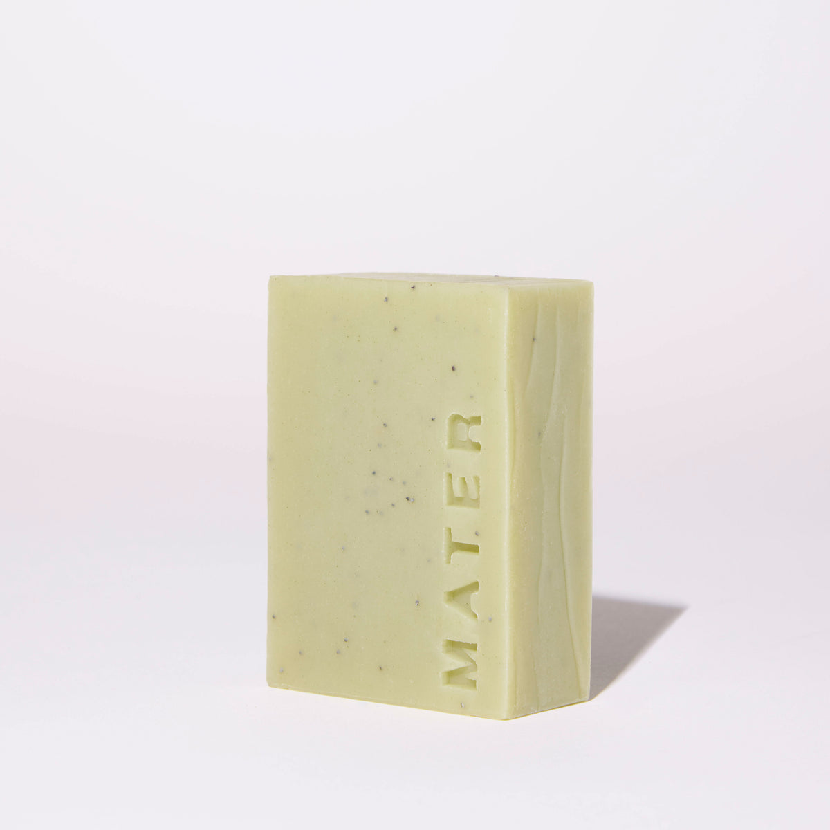 A rectangular bar of MATER soap in a pale sage green color with tiny dark specks, standing upright on a white surface against a white background.