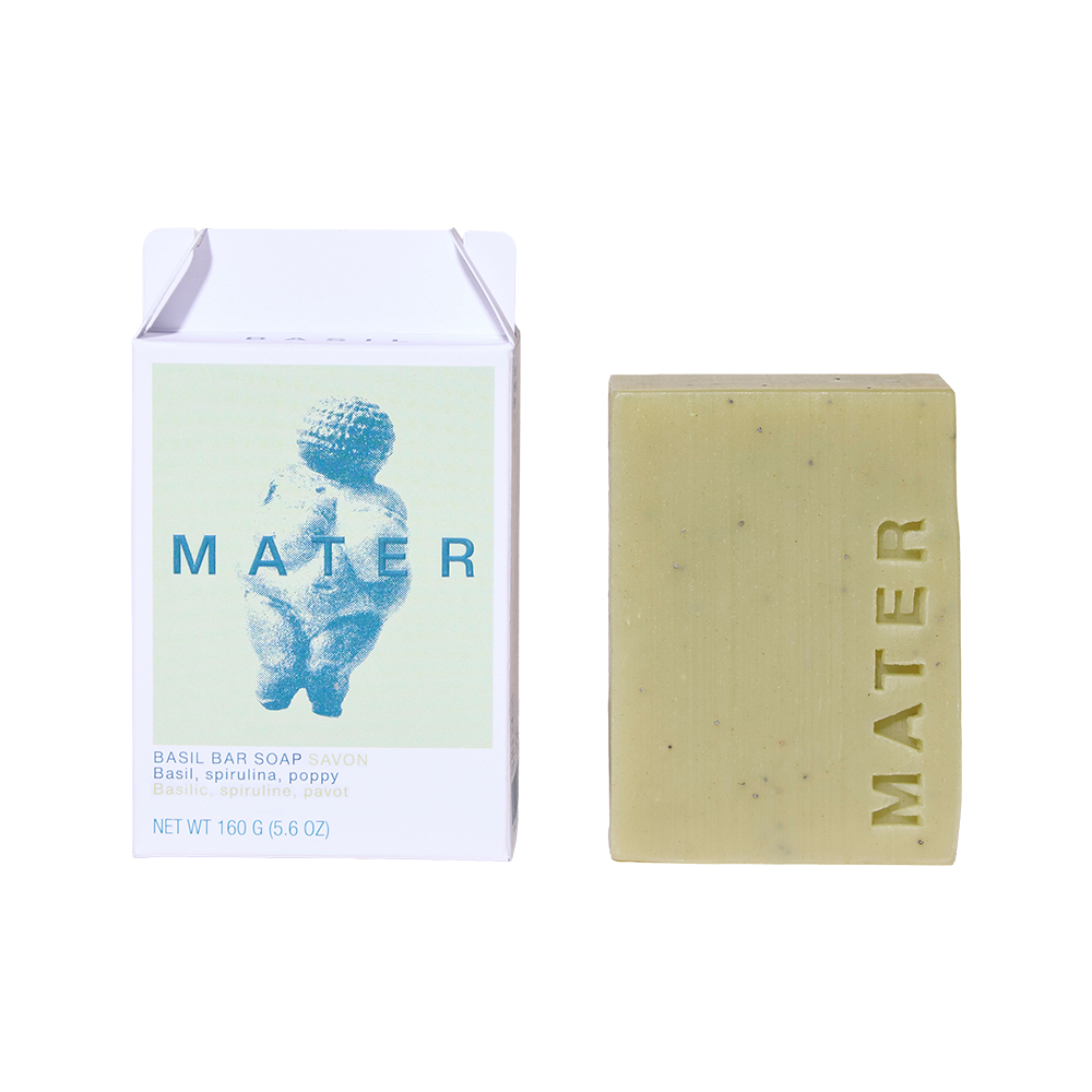 A bar of MATER basil bar soap next to its white cardboard packaging box, which features a light blue illustration of the Bellerose baby statue.