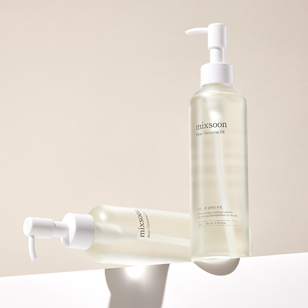 Two clear pump bottles of mixsoon Bean Cleansing Oil with white caps, one standing upright and one lying horizontally, on a clean beige background with soft shadows.