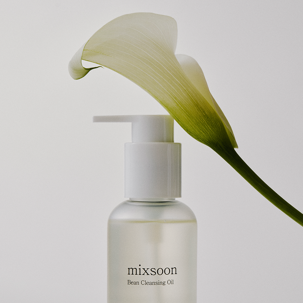 A bottle of mixsoon Bean Cleansing Oil with a white pump dispenser, styled with a white calla lily flower on a light gray background.