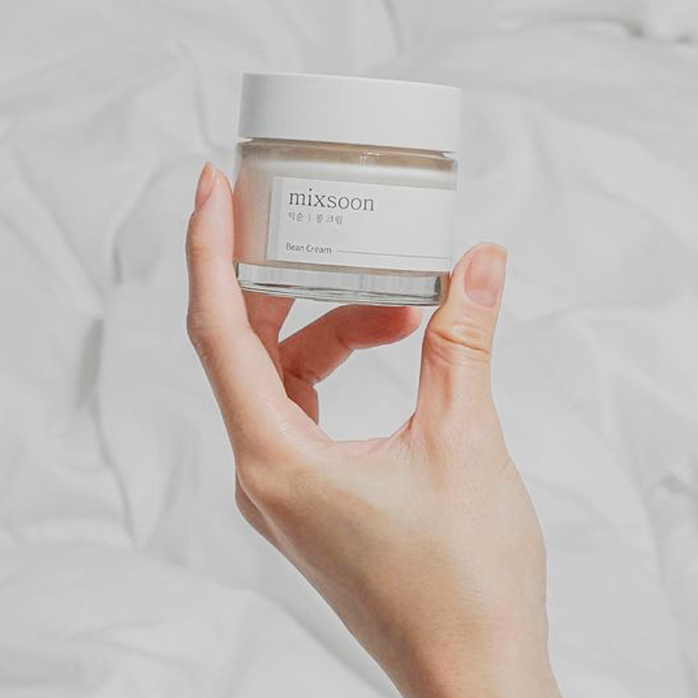 A hand holding a small clear jar of Mixsoon Bean Cream against a soft, white wrinkled fabric background.