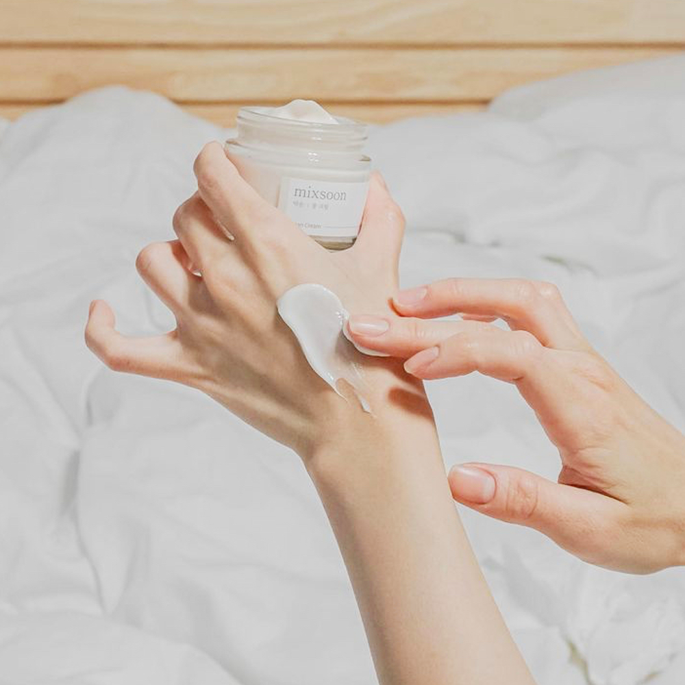 A person applies Mixsoon Bean Cream onto their hand, set against a soft, white bedding background with a light wood headboard.