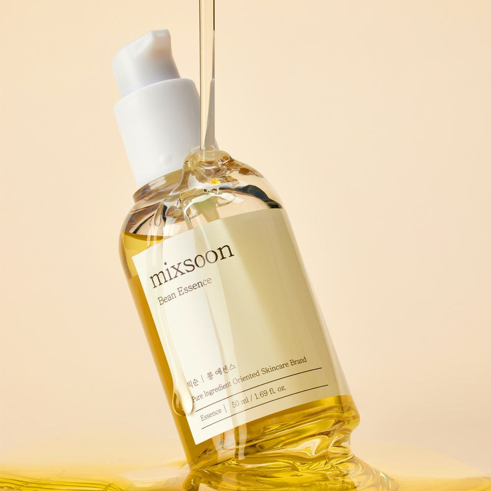 A bottle of mixsoon Brain Essential oil serum with a white pump dispenser, filled with golden yellow oil, set against a warm beige background with a drop of oil dripping into it.