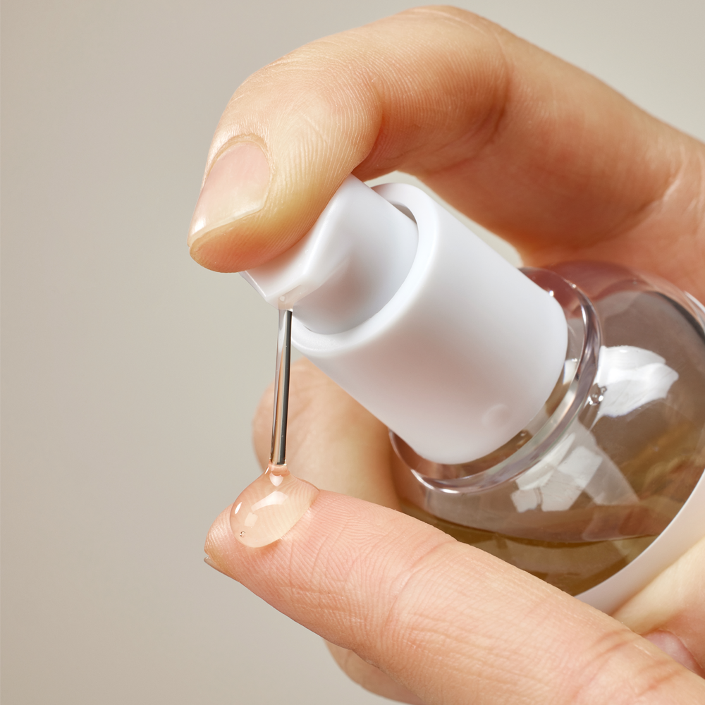 A close-up of a hand pressing the white pump of a clear cosmetic bottle, with a small drop of pale pink liquid resting on the fingertip.