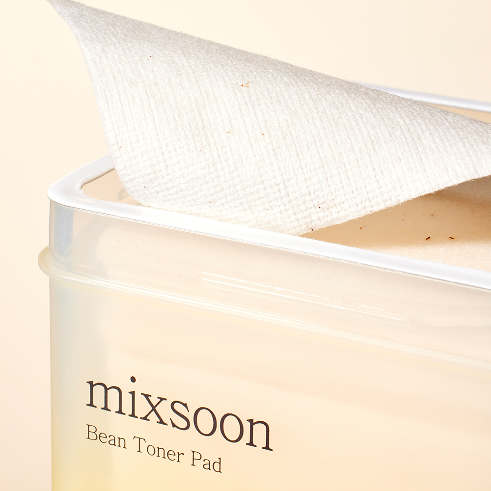 Close-up of a mixsoon Bean Toner Pad being pulled from its clear plastic container, showing the textured white cotton pad and pale beige toner inside.