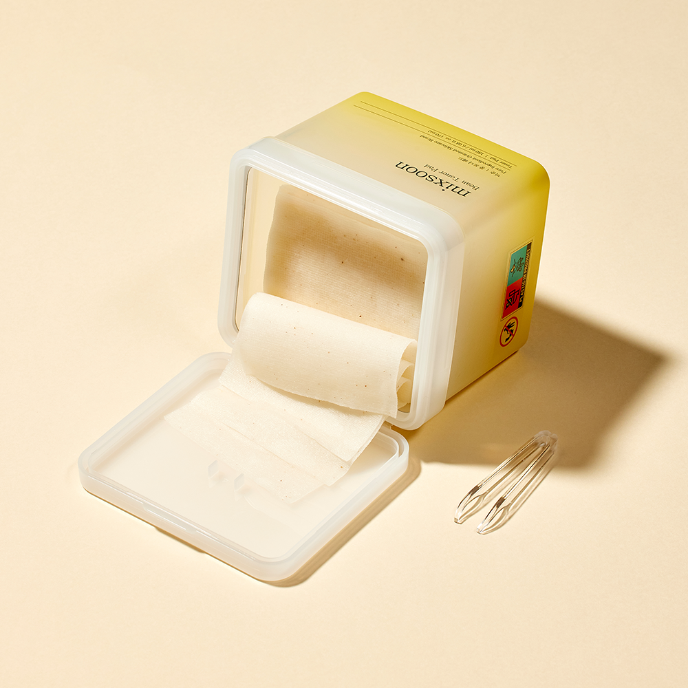mixsoon Bean Toner Pad container with a clear lid, shown open with individual pads folding out of it and small plastic tongs to the side on a warm beige background. 