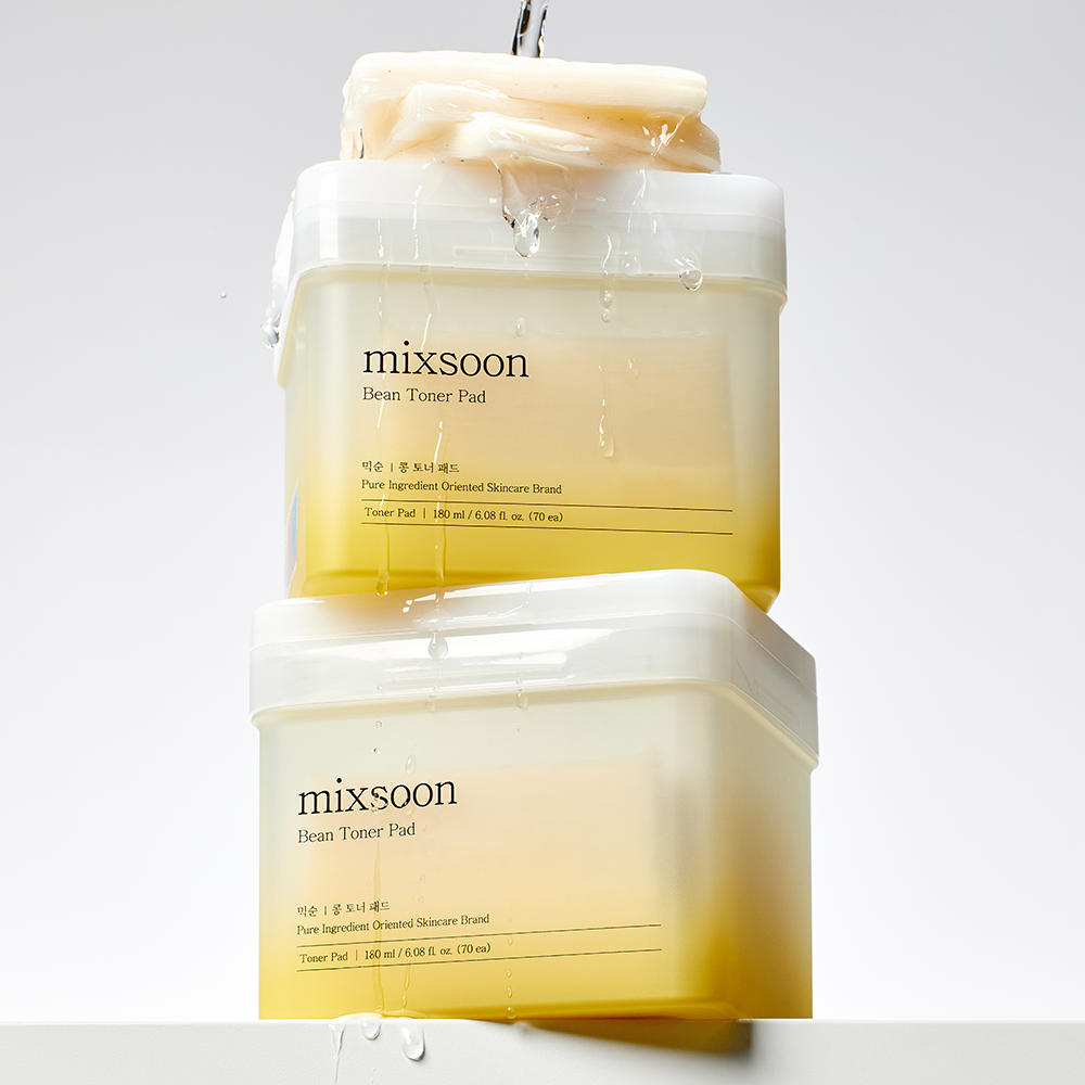 Two containers of mixsoon Bean Toner Pad stacked vertically, with water pouring on them. The toner pads show a yellow-to-cream gradient inside translucent white jars against a light gray background.