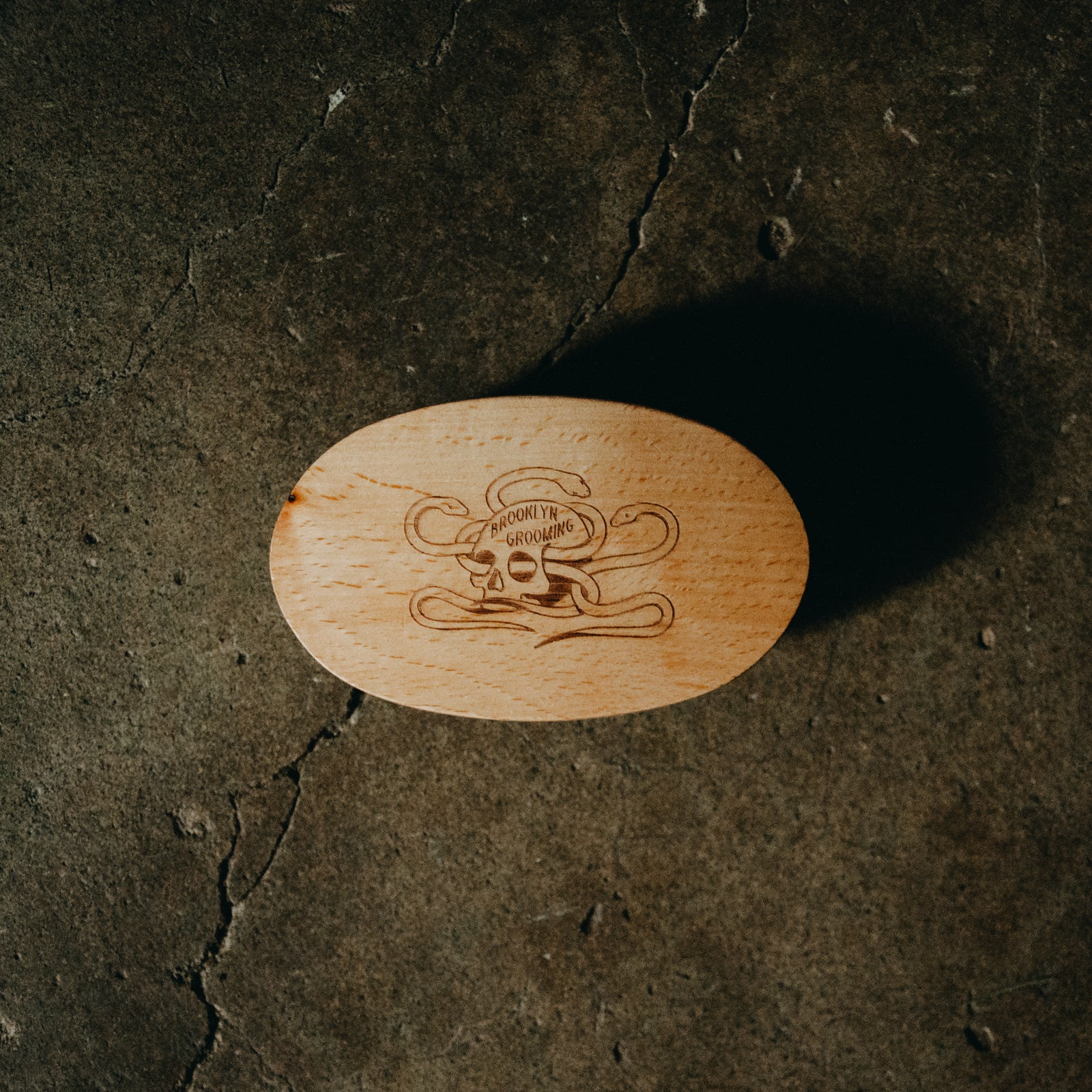An oval beard brush with a carved skull and crossbones design and the words 'Brooklyn Grooming' on a dark textured concrete surface.