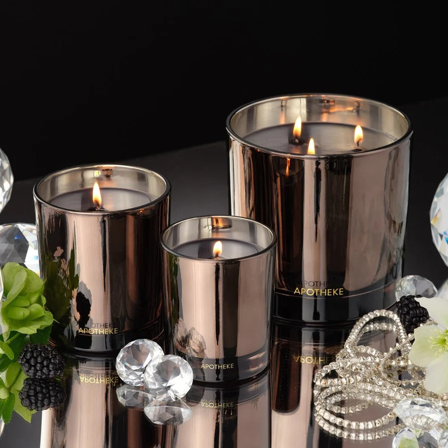 Three Apotheke candles in metallic containers on a reflective surface with decorative elements.