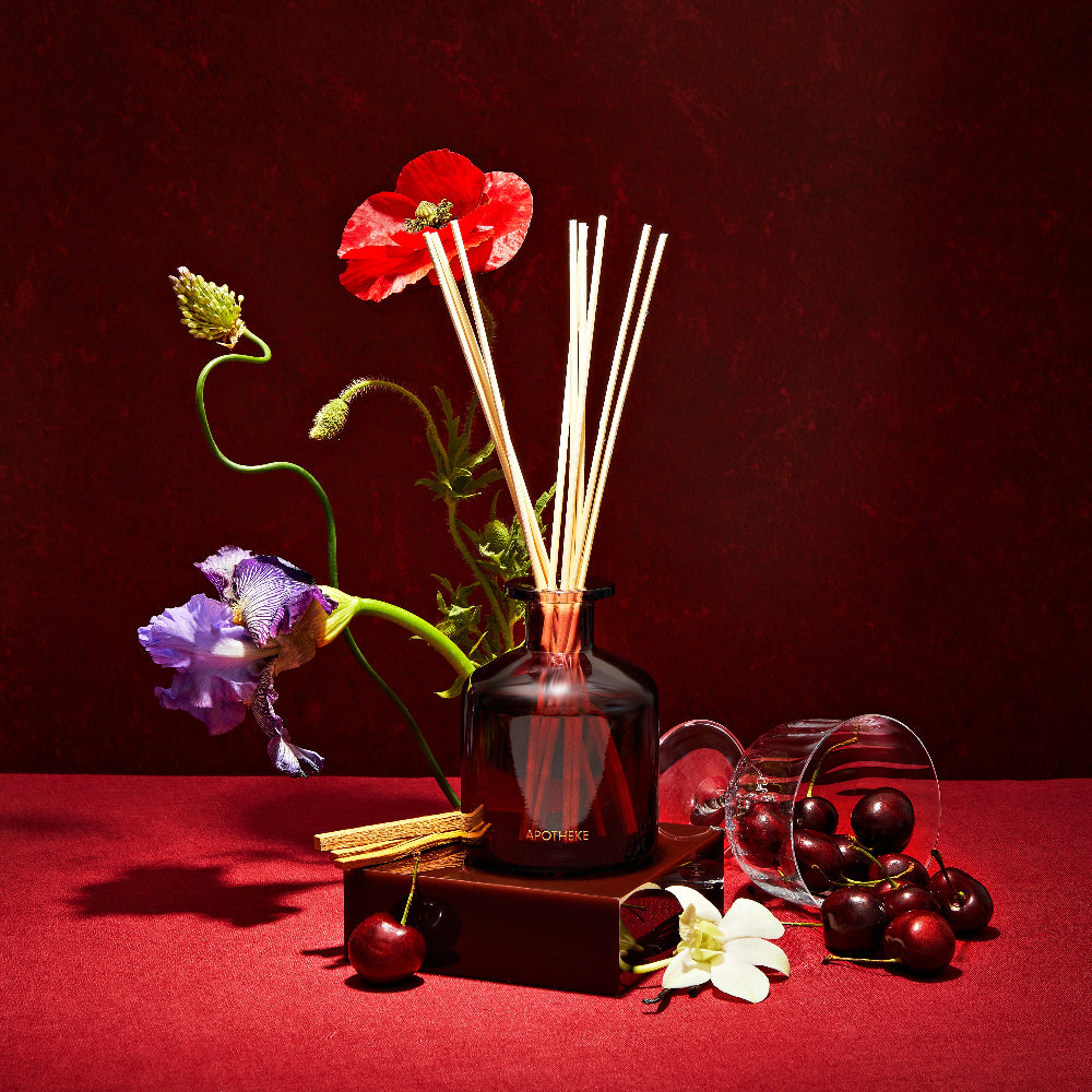 Apotheke Bitter Cherry Diffuser with floral reeds, flowers, and cherries on a red surface with a dark background