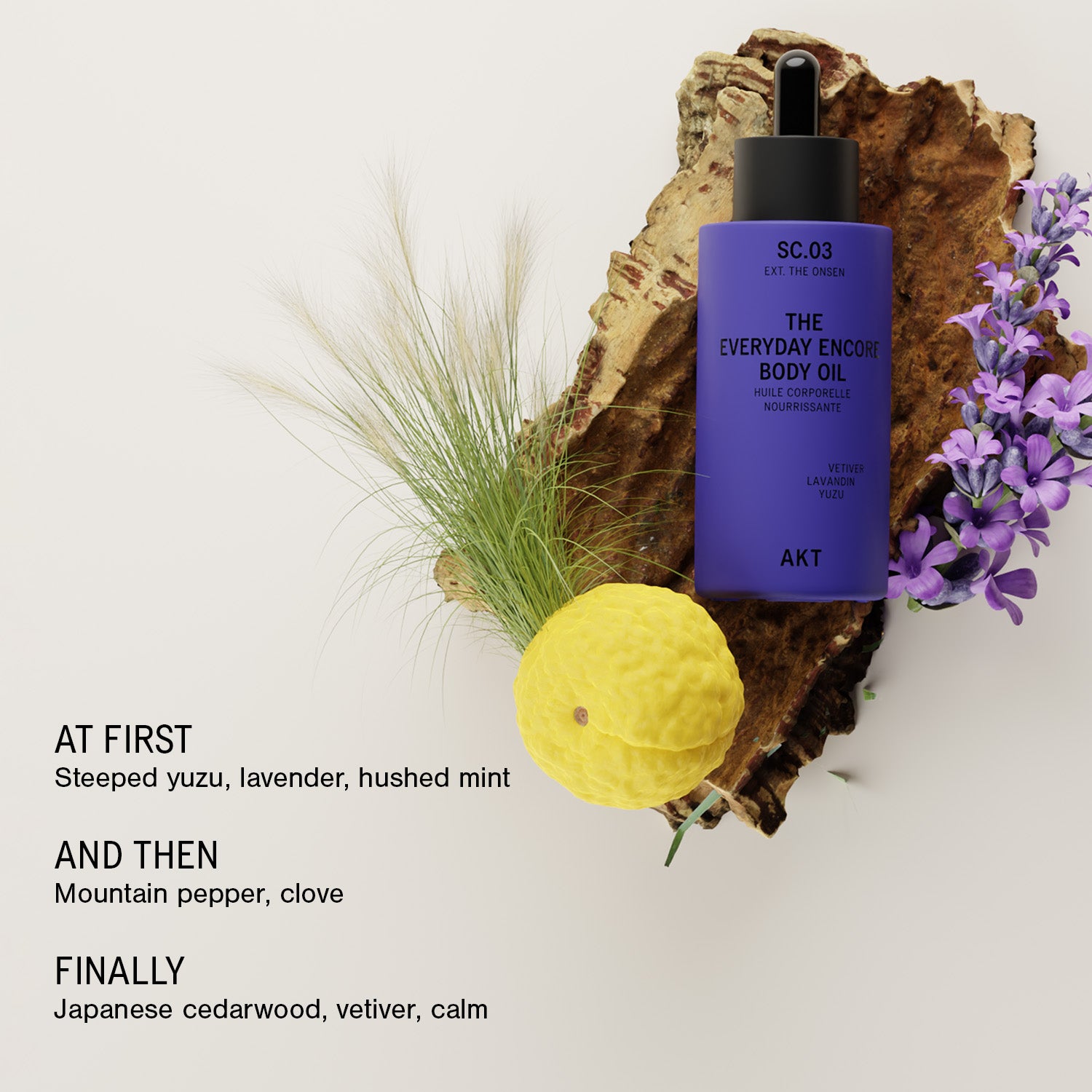 A bottle of AKT The Everyday Encore Body Oil Sc.03 stands on a piece of wood with decorative green grass, purple flowers, and a yellow citrus slice against a light neutral background. Text on the left reads: 'AT FIRST Steeped yuzu, lavender, hushed mint AND THEN Mountain pepper, clove FINALLY Japanese cedarwood, vetiver, calm.