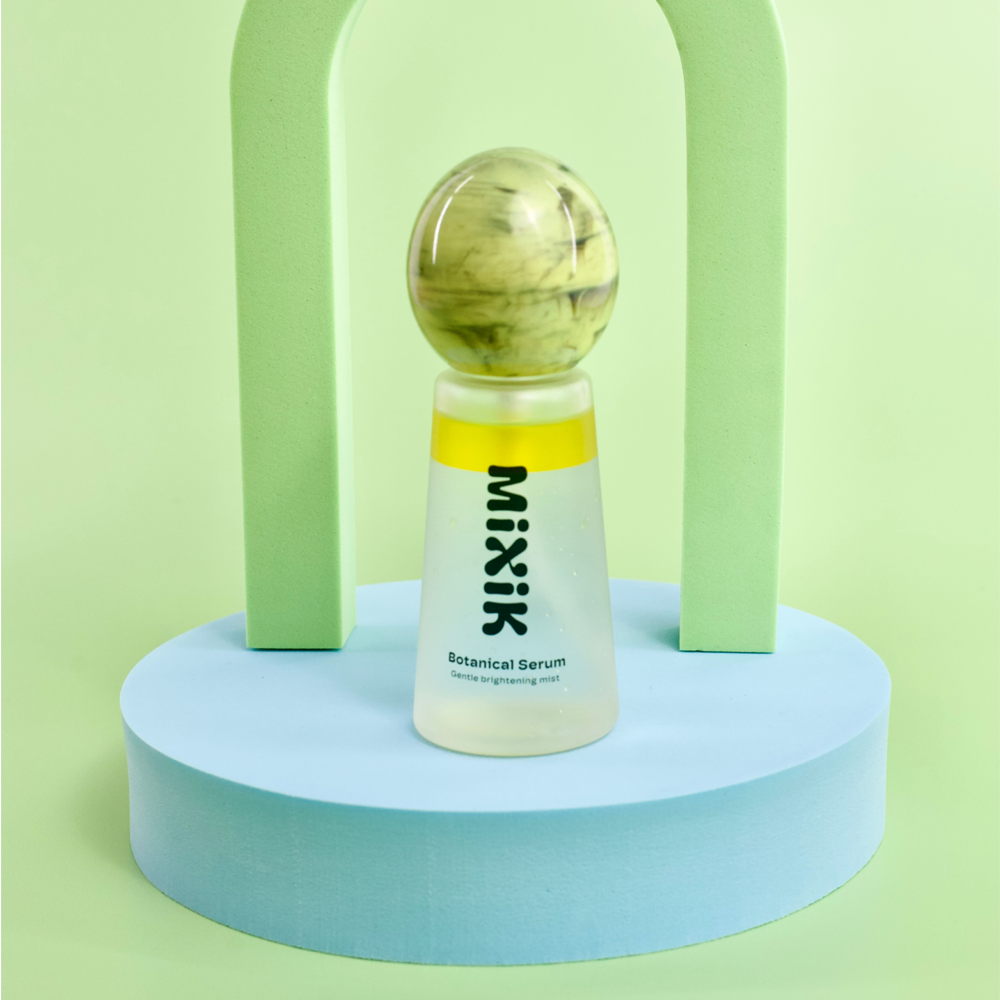 A bottle of Mixik Botanical Serum stands centered on a light blue circular pedestal, with two pale green arch shapes behind it against a soft mint-green background.