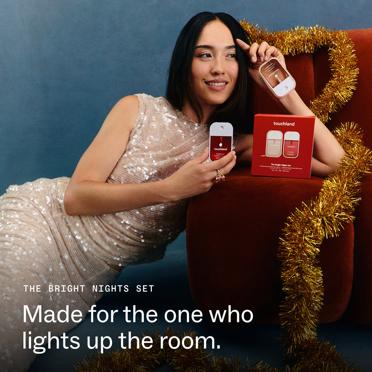 A woman in a sequined champagne dress reclines on a red armchair wrapped with gold tinsel, holding a Touchland light-up night light and its red box. Text on the image reads 'THE BRIGHT NIGHTS SET' and 'Made for the one who lights up the room.