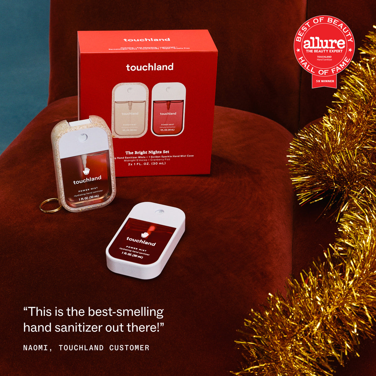 Touchland The Bright Night Set hand sanitizer and sanitizer keychain set displayed on a dark red velvet chair next to its red packaging box, with a gold tinsel decoration and an Allure Best of Beauty Mill of Fair award badge.