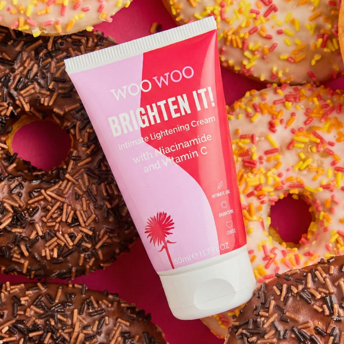 A squeeze tube of Woo Woo Brighten It! Intimate Lightening Cream is placed diagonally across a pile of decorated donuts. The tube is pink and red with white text, and the background is filled with donuts topped with colorful sprinkles.