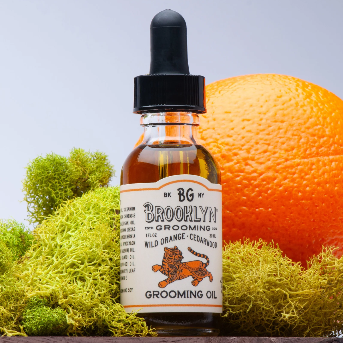 Bottle of Brooklyn Grooming oil with a dropper on a wooden surface with an orange and greenery in the background.