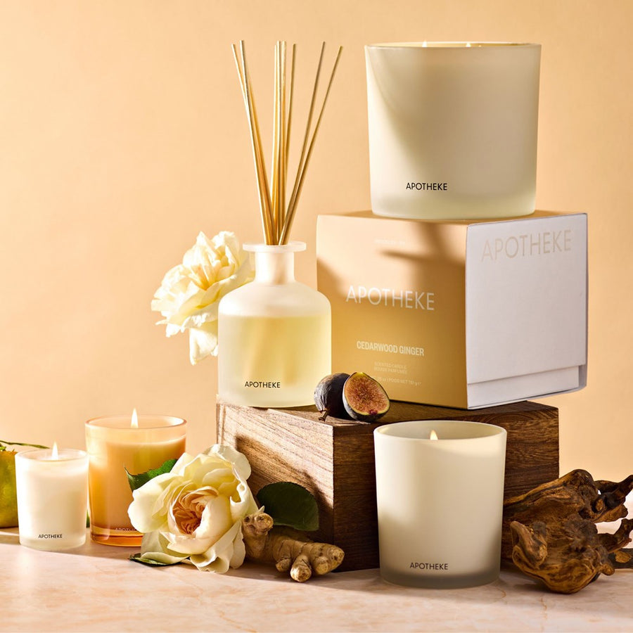 A styled product arrangement of APOTHEKE candles and a reed diffuser on a warm beige background. The set includes a large frosted glass jar candle, a smaller white glass jar candle, a reed diffuser bottle with wooden sticks, a cream-colored box, and additional candles and botanical props on a wooden block.