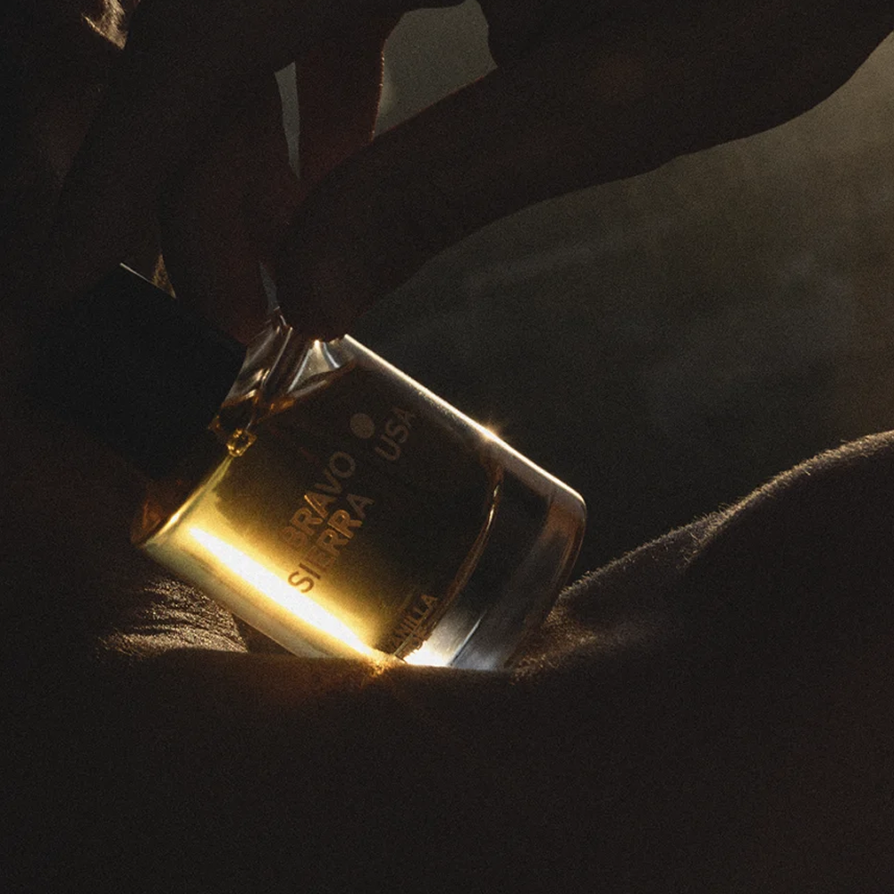 A close-up of a hand holding a bottle of Bravo Sierra Vanilla Hide - Eau de Parfum  which glows with a warm golden-yellow light against a dark, shadowed background.