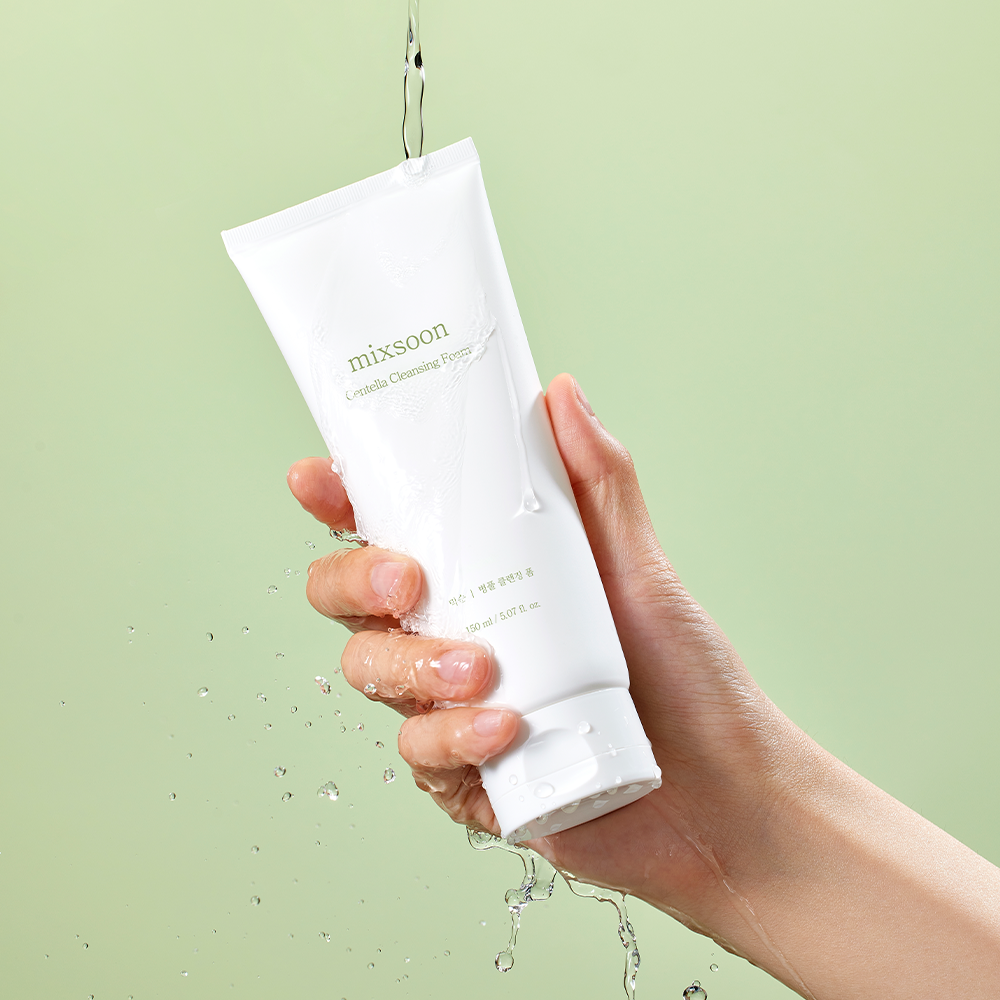 A hand holding a white tube of Mixsoon Centella Cleansing Foam under running water against a light green background.