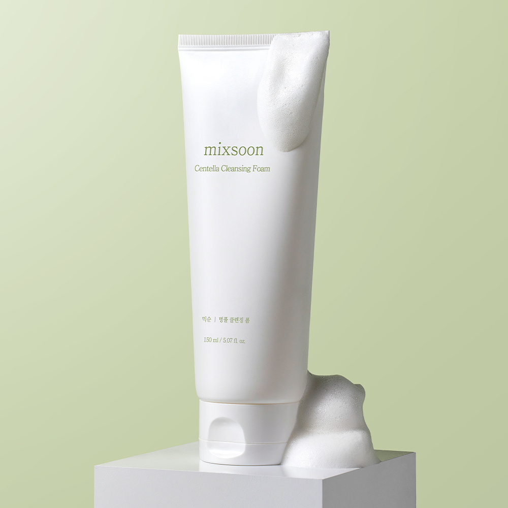 A white squeeze tube of Mixsoon Centella Cleansing Foam standing upright on a white pedestal against a pale green background, with foam dripping from the top and resting on the base.