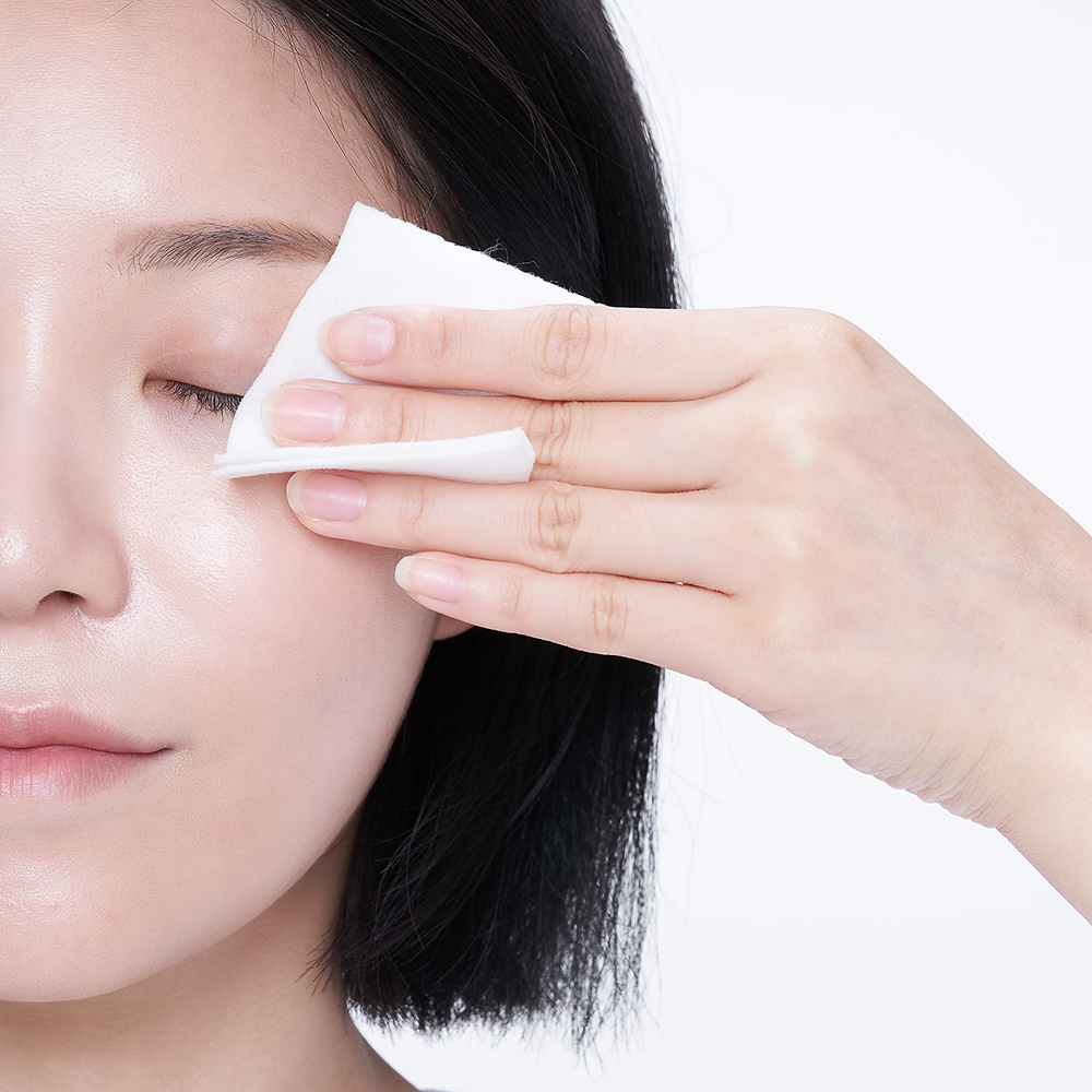 A close-up of a person with dark hair using mixsoon Centella Cleansing Water to gently wipe their eye area against a clean white background.
