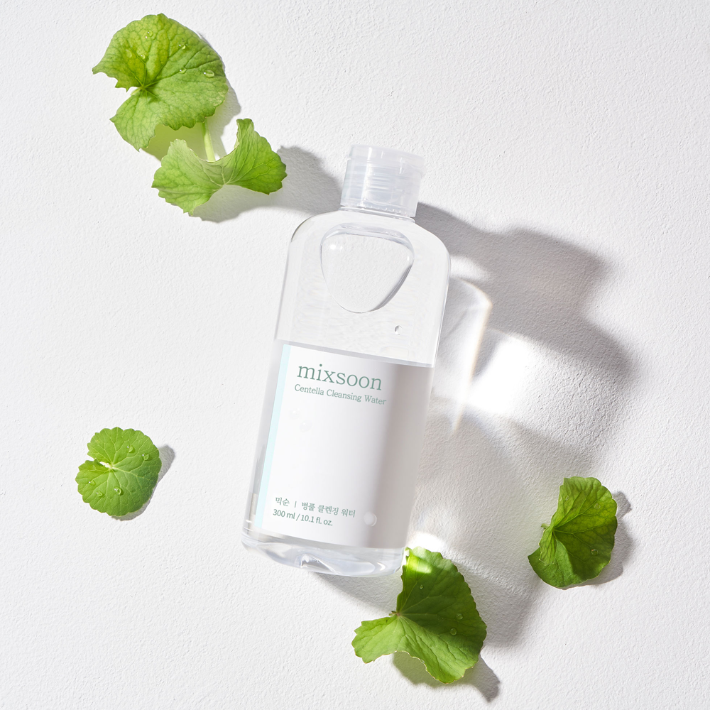 A clear plastic bottle of mixsoon Centella Cleansing Water with a white label, styled on a white textured background with green leaves scattered around it.