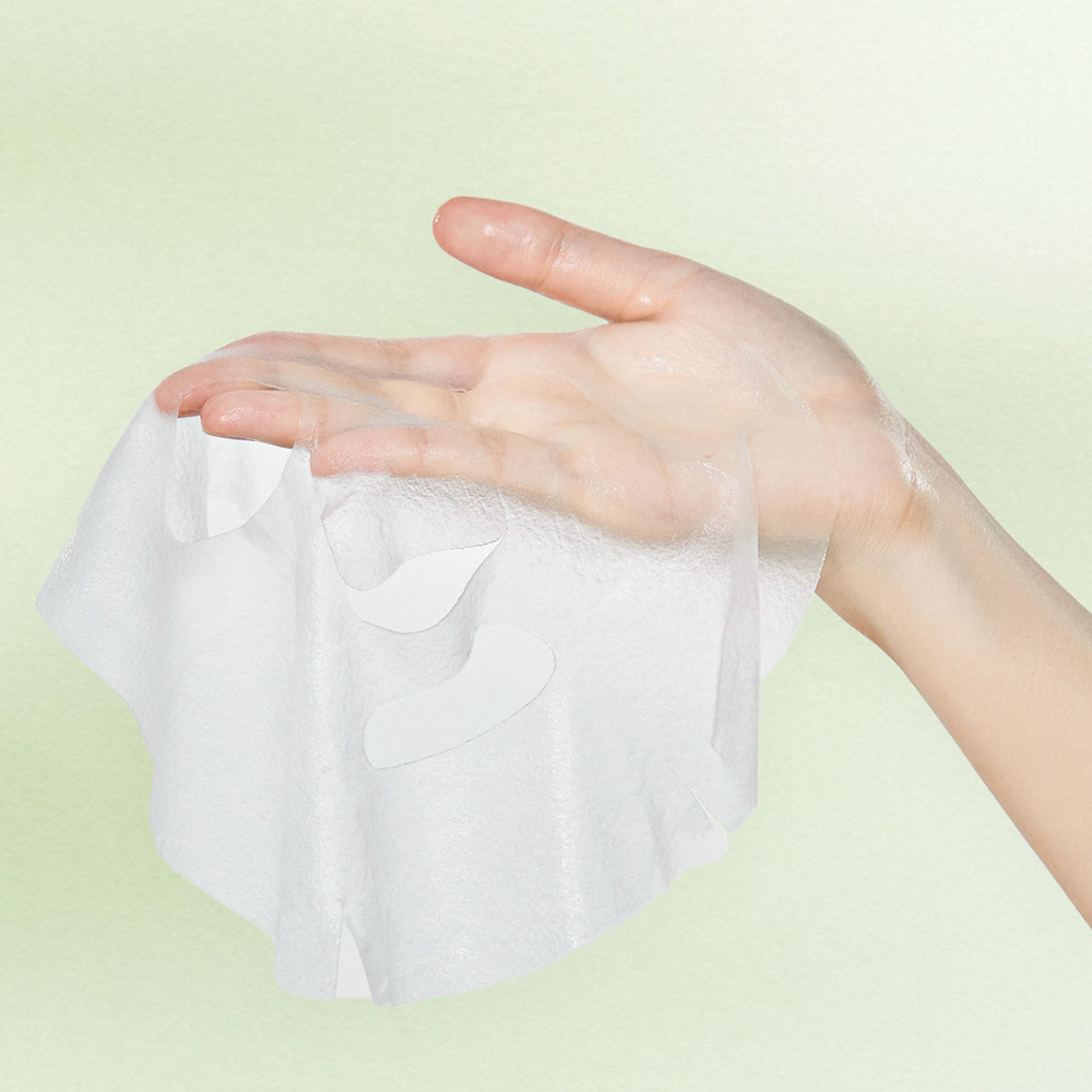 A translucent white sheet mask being held in a person's hand against a soft pale green background.