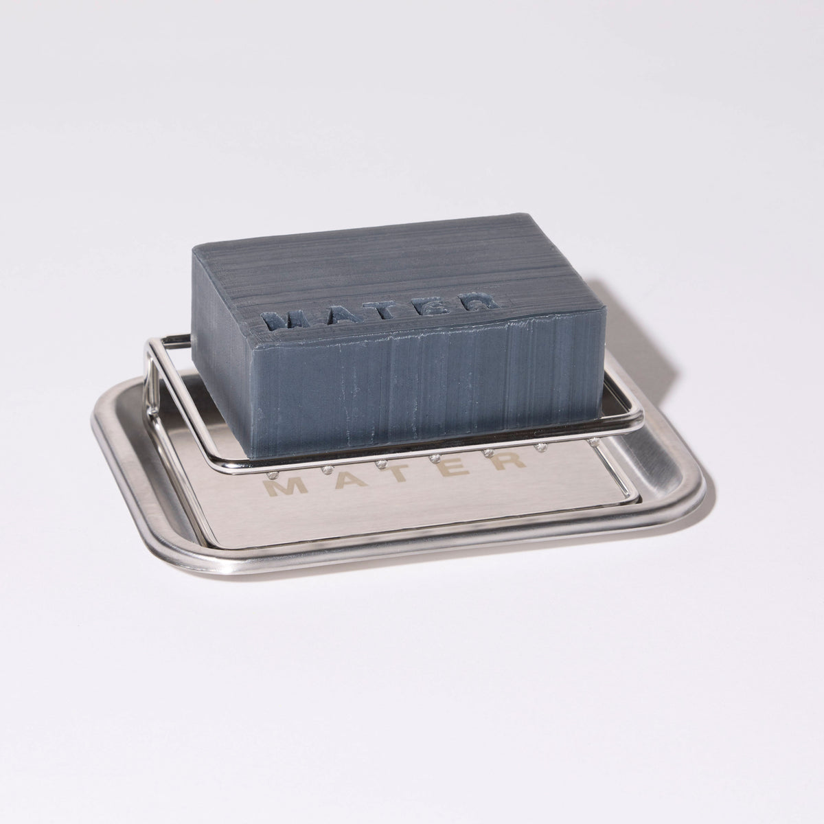A dark slate blue soap bar with 'MATER' embossed on top, resting on a brushed silver metal soap dish also engraved with 'MATER'.
