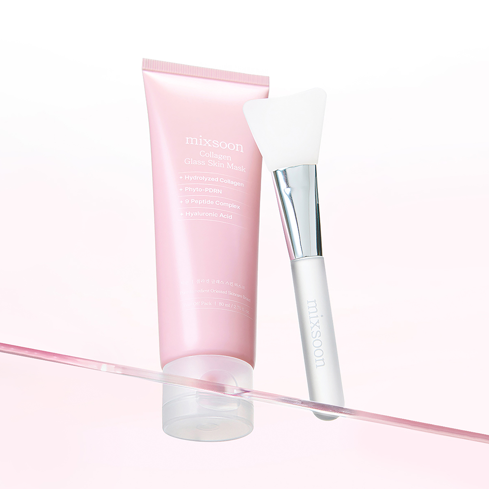 A pink Mixsoon Collagen Clear Skin Mask tube standing next to a white and silver makeup brush on a reflective pink surface against a white background.