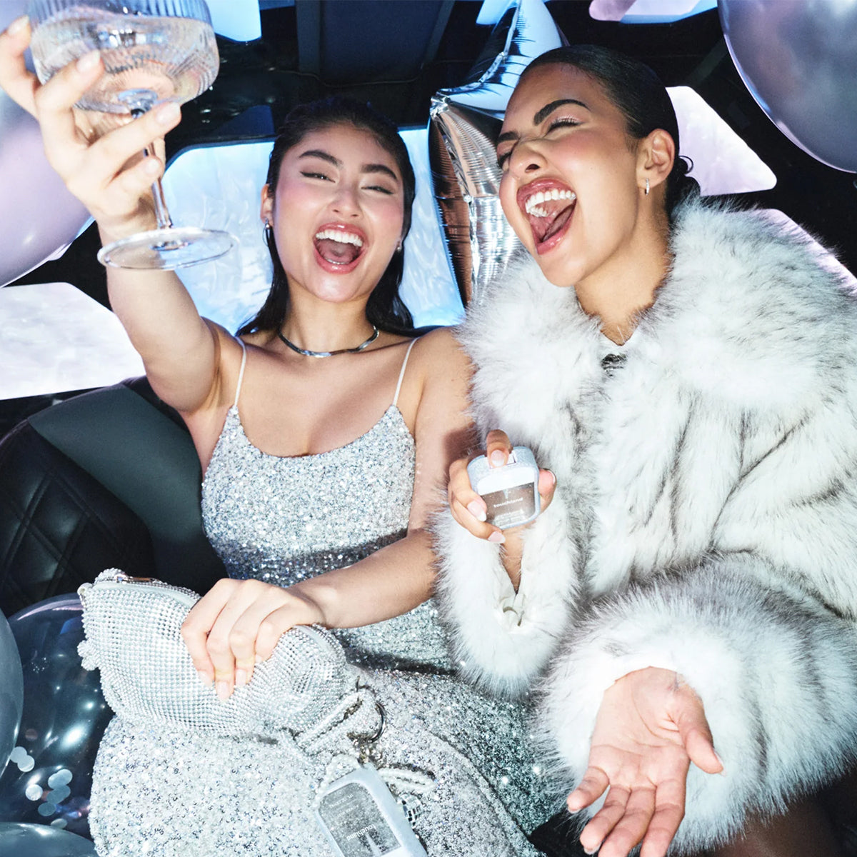 Two women laughing and celebrating inside a car, one wearing a sparkling silver dress and holding a glass, and the other wearing a white fur coat while holding Touchland Power Mist Hydrating Hand Sanitizer - Confetti Cake.