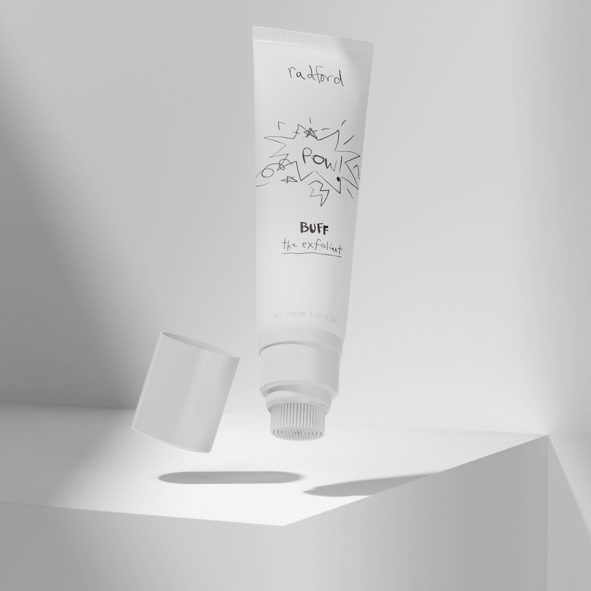 Tube of Radford Buff the Exfoliant floating above a white geometric platform, with its cap detached and lying to the left. The scene is rendered in soft gray and white tones with clean studio-style shadows.