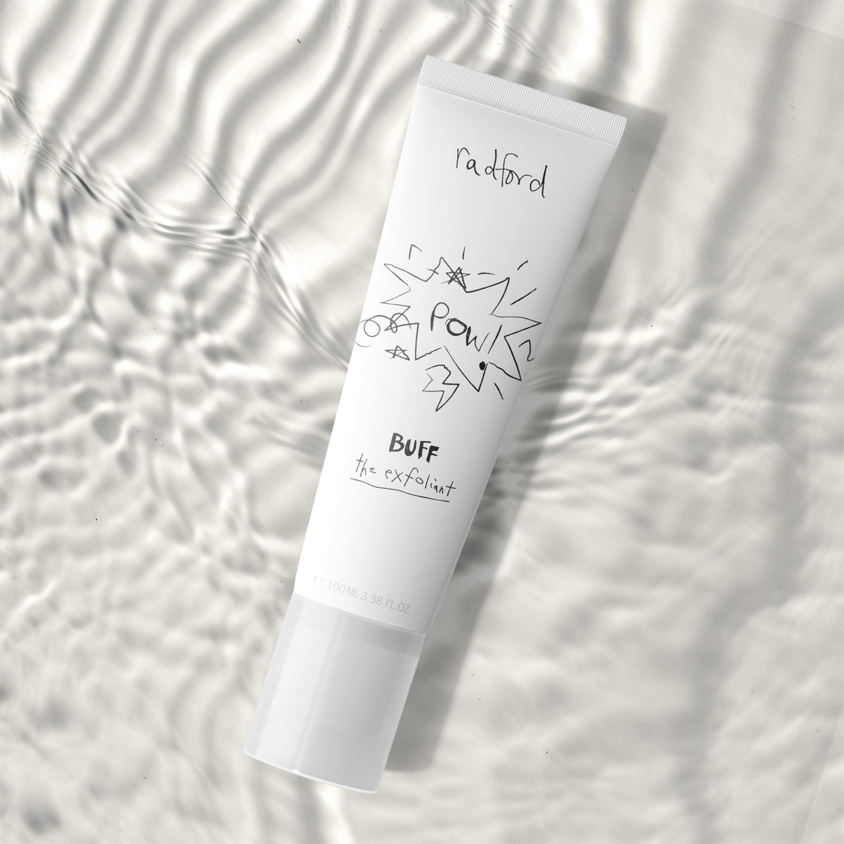 A white squeeze tube of Radford Buff the Exfoliant, featuring black handwritten-style text and graphics, placed diagonally on a white textured surface with soft wavy shadows.