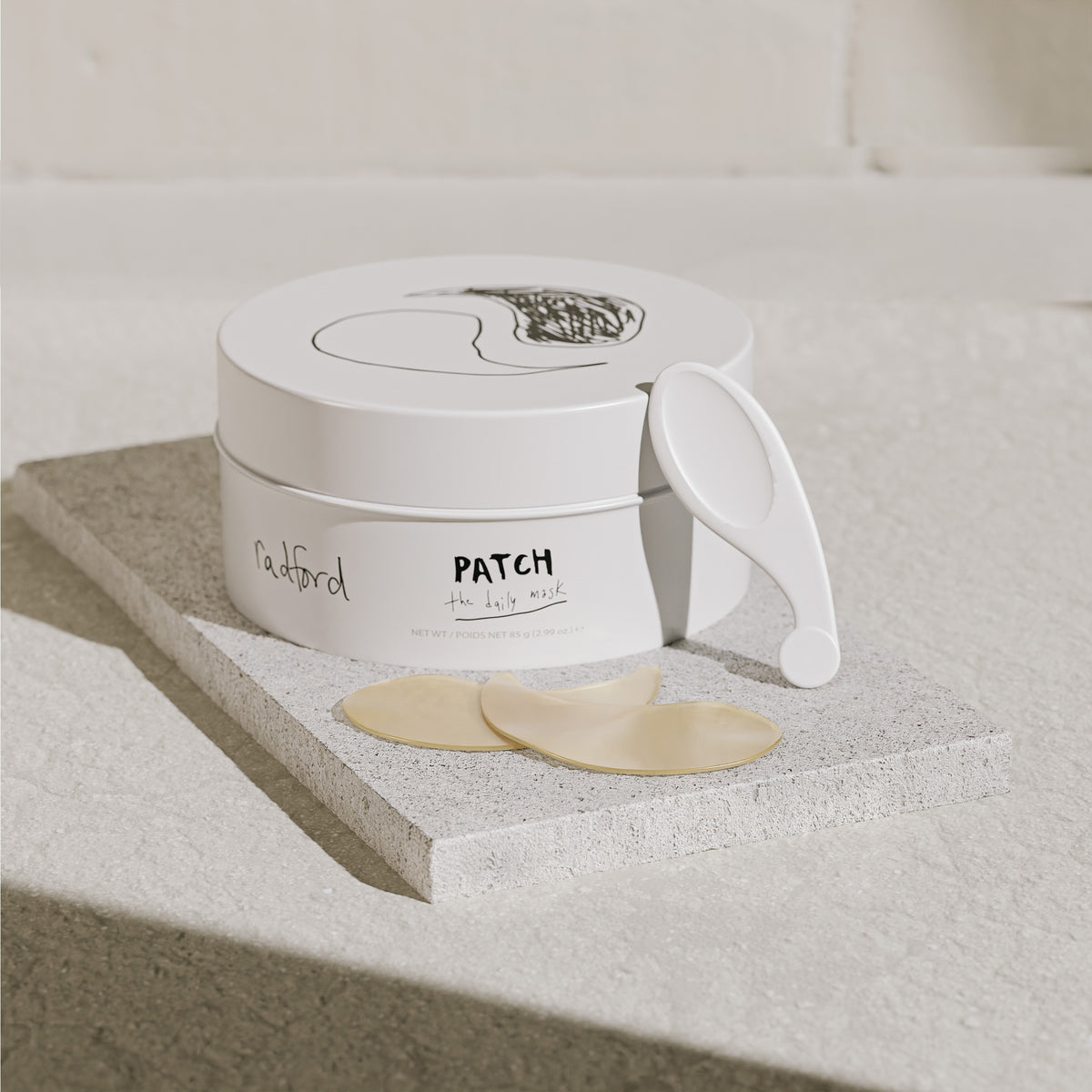 A white jar of Radford Patch - the daily mask with a white applicator spoon resting beside it, set on a textured stone slab with two beige mask patches in front. The background is a clean, neutral beige stone surface.
