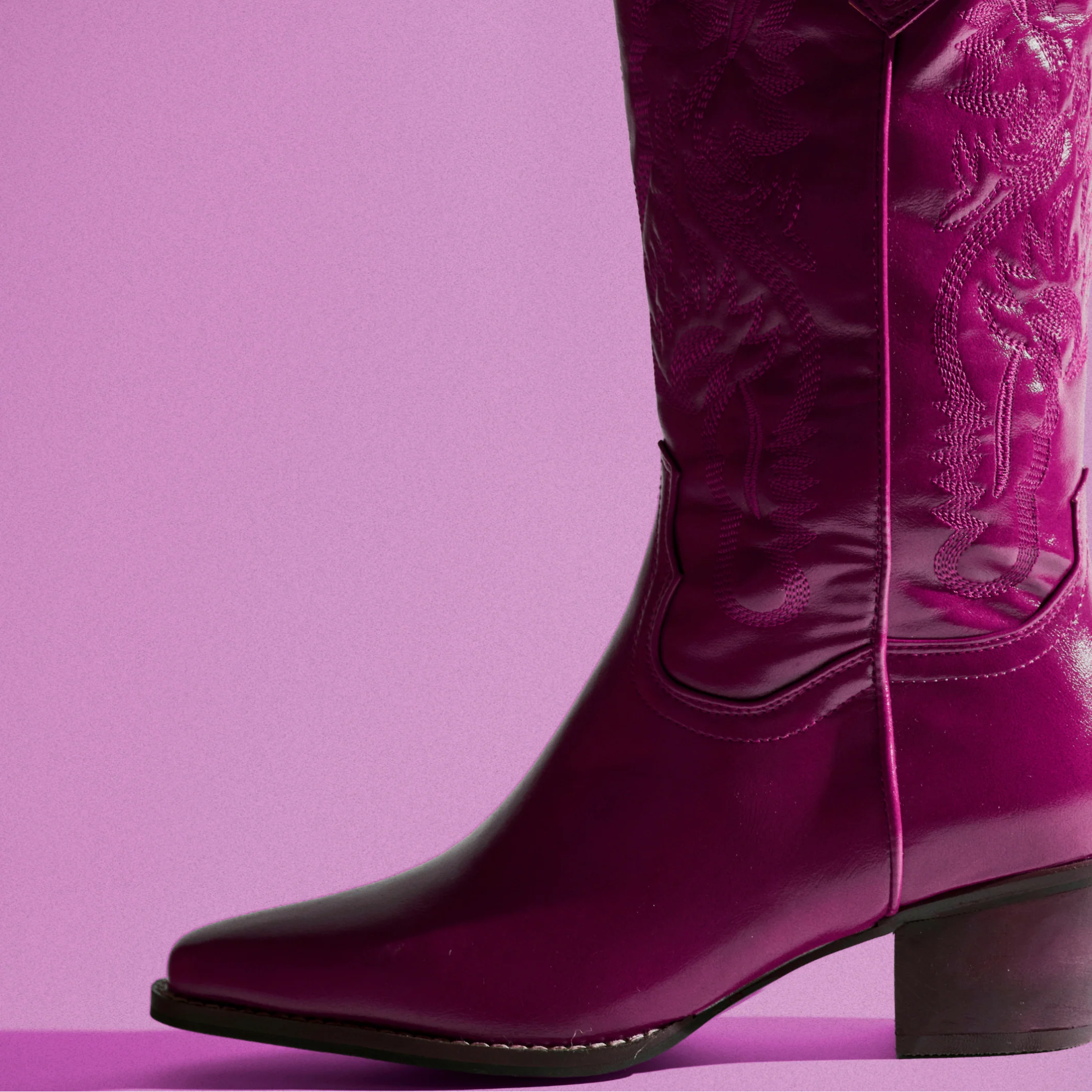 A single glossy magenta western boot with embossed floral detailing on the shaft, shown against a solid lavender-pink background.