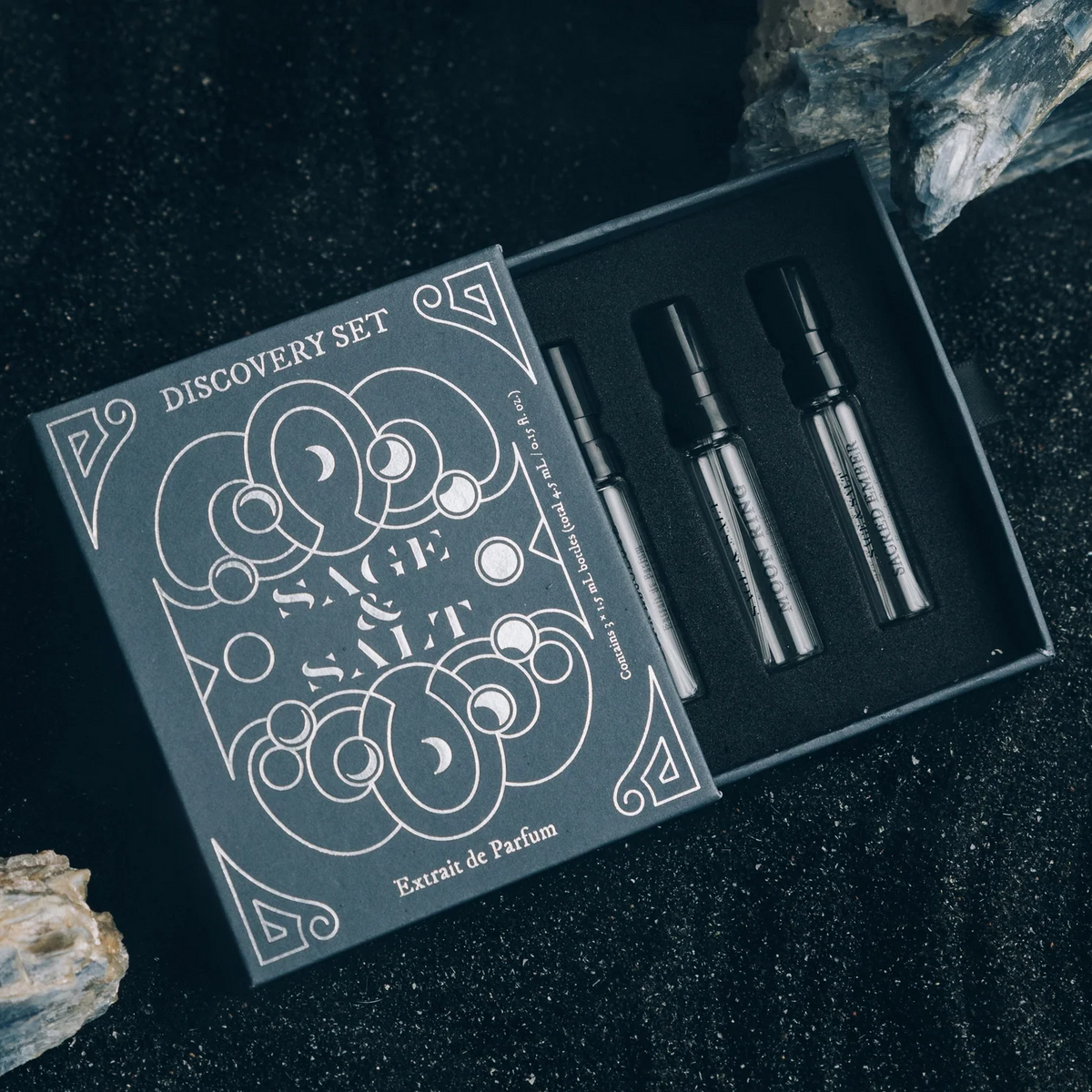 A boxed fragrance discovery set labeled 'Sage Salt' with three dark glass sample vials placed in a black tray beside the box, styled on a dark textured surface with driftwood.