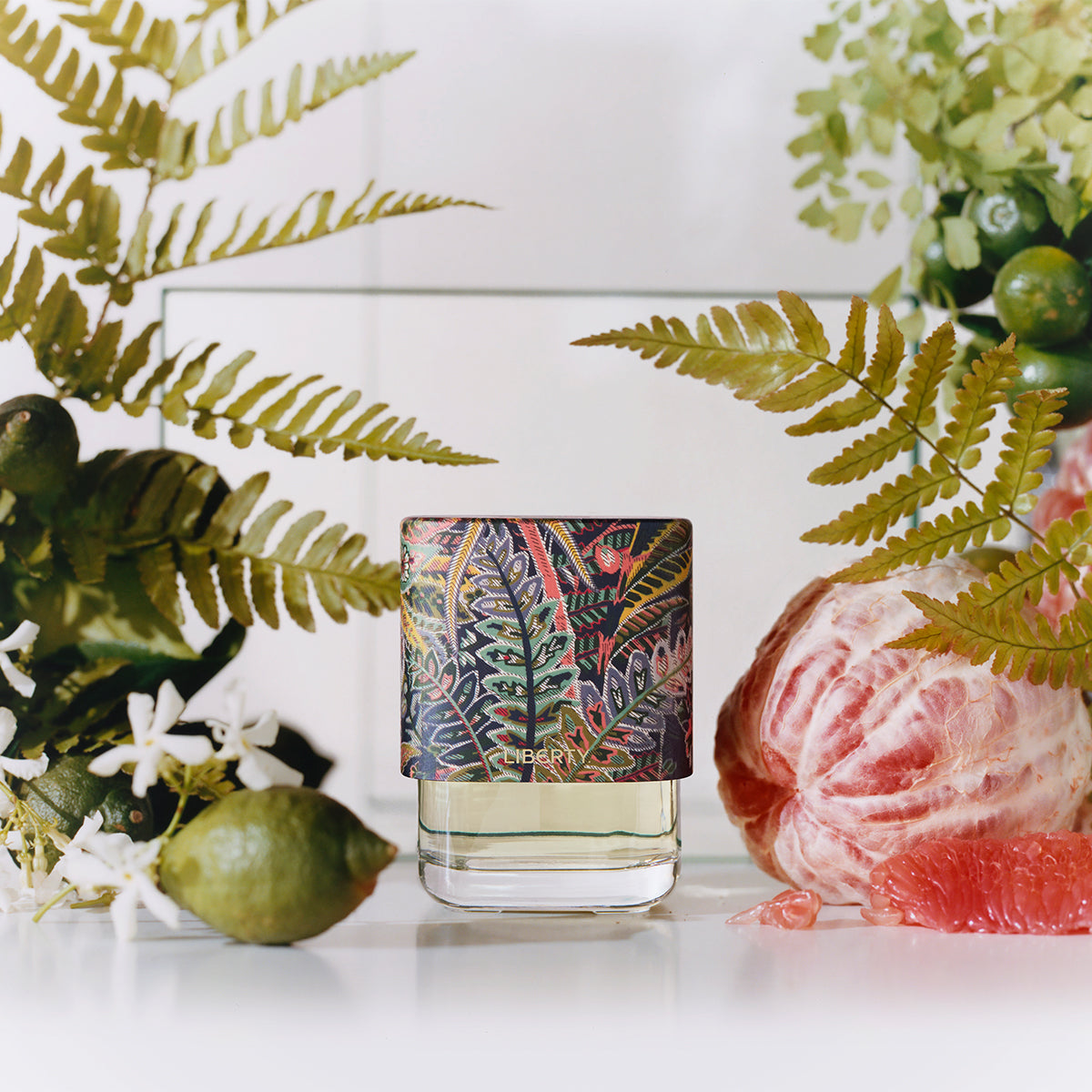 Perfume bottle with a decorative label surrounded by greenery and fruit on a white surface