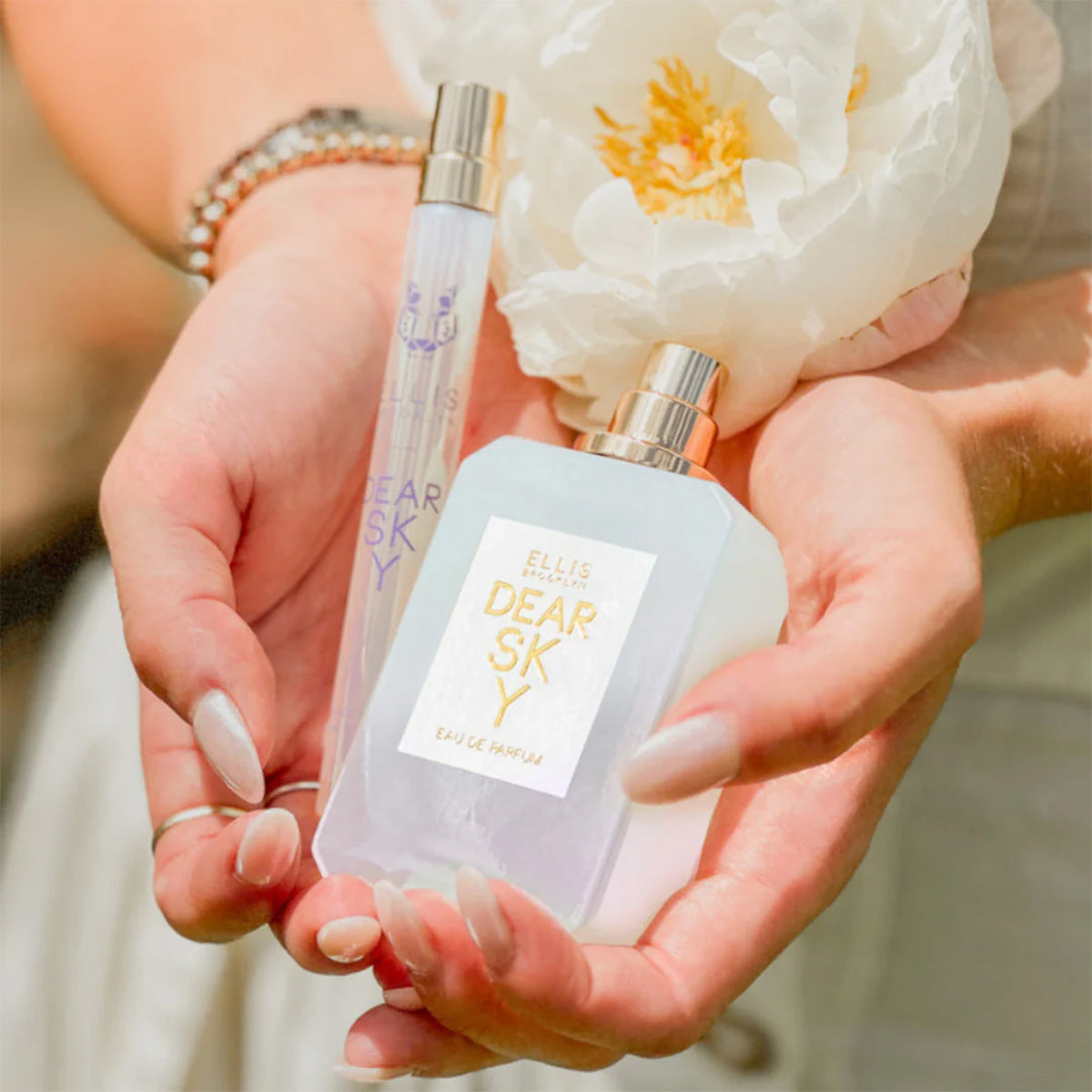 Hands holding Dear Sky full size perfume and travel spry in hands with a flower.
