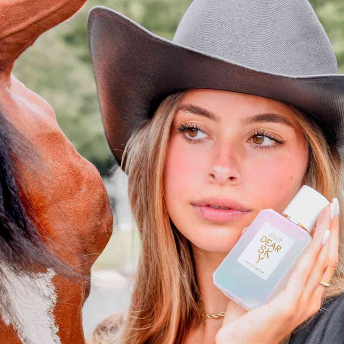 Woman wearing a cowboy hat next to a horse holding dear sky eau de parfum