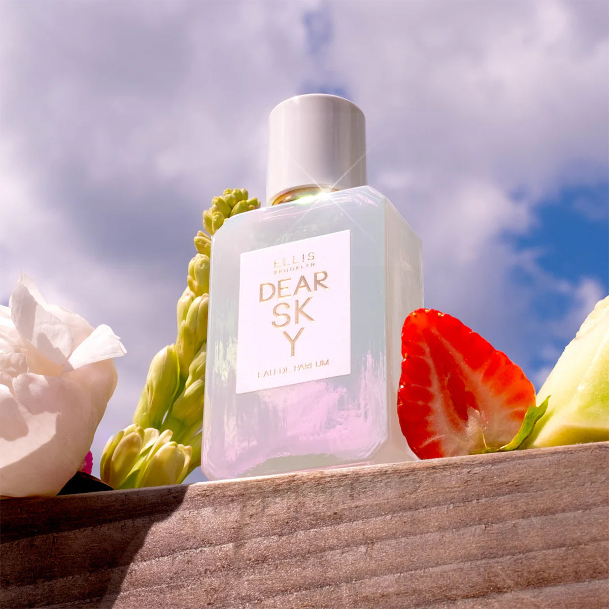 Dear sky eau de parfum on a wood ledge next to flowers and fruit in front of blue sky and clouds.
