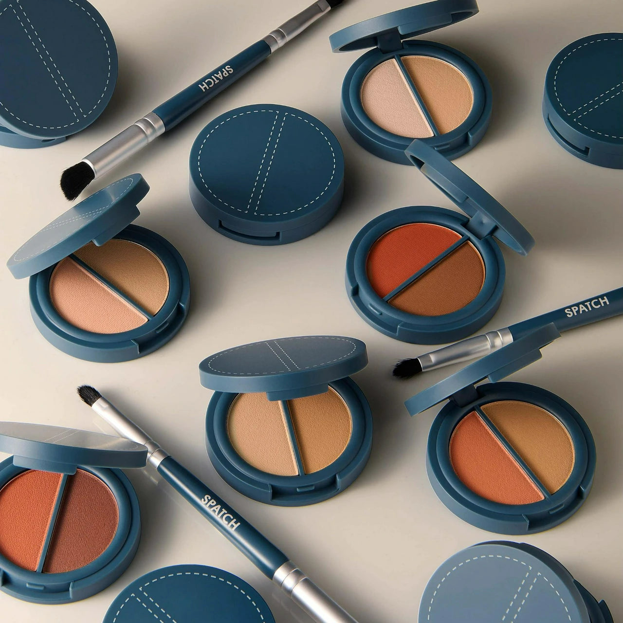 A styled flat-lay of round makeup compacts and dual-ended brushes with blue-gray casing and silver handles, arranged on a light beige background. The compacts are open to reveal two-toned face powders in warm beige, tan, peach, and orange shades.