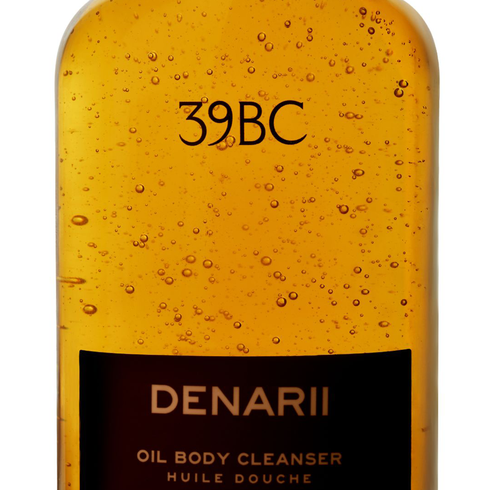 A close-up of a 39BC Denarii Oil Body Cleanser bottle showing the golden-yellow translucent oil inside, with visible droplets on the surface. The label features a black block at the bottom with the brand name 'DENARII' and the text 'OIL BODY CLEANSER'.