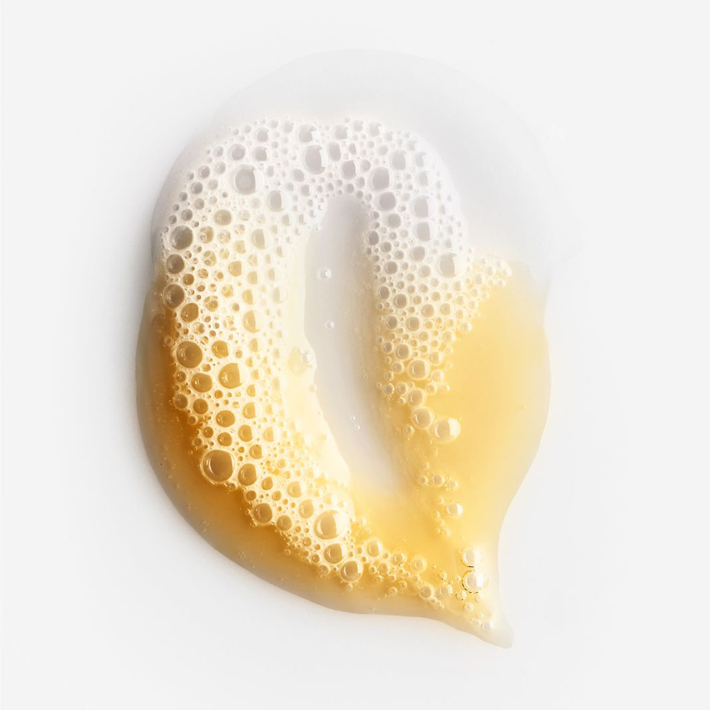 A glossy, translucent droplet of pale golden-yellow liquid with dense white foam bubbles, photographed on a clean white background.