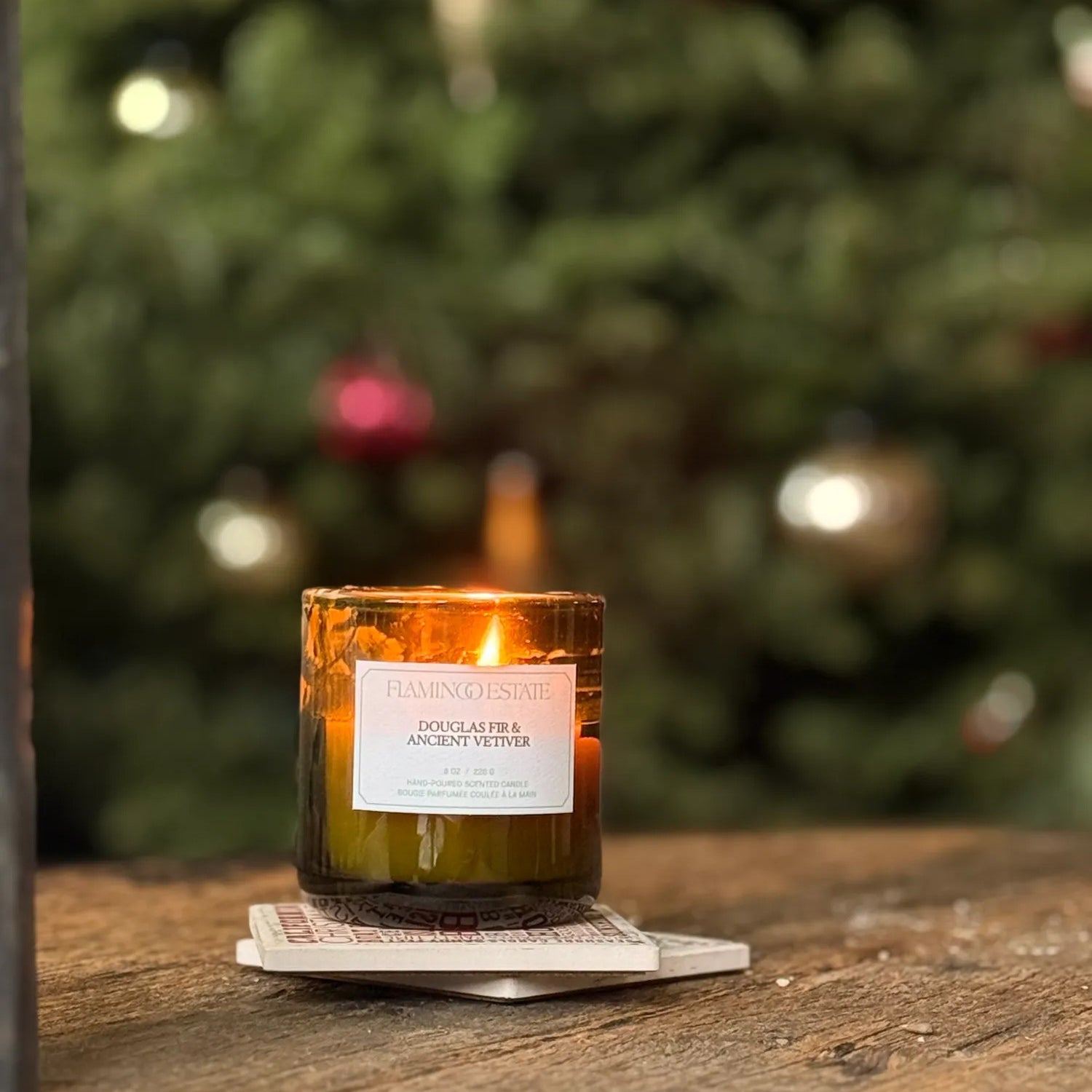Flamingo Estate Candle with a label on a coaster in front of a blurred Christmas tree