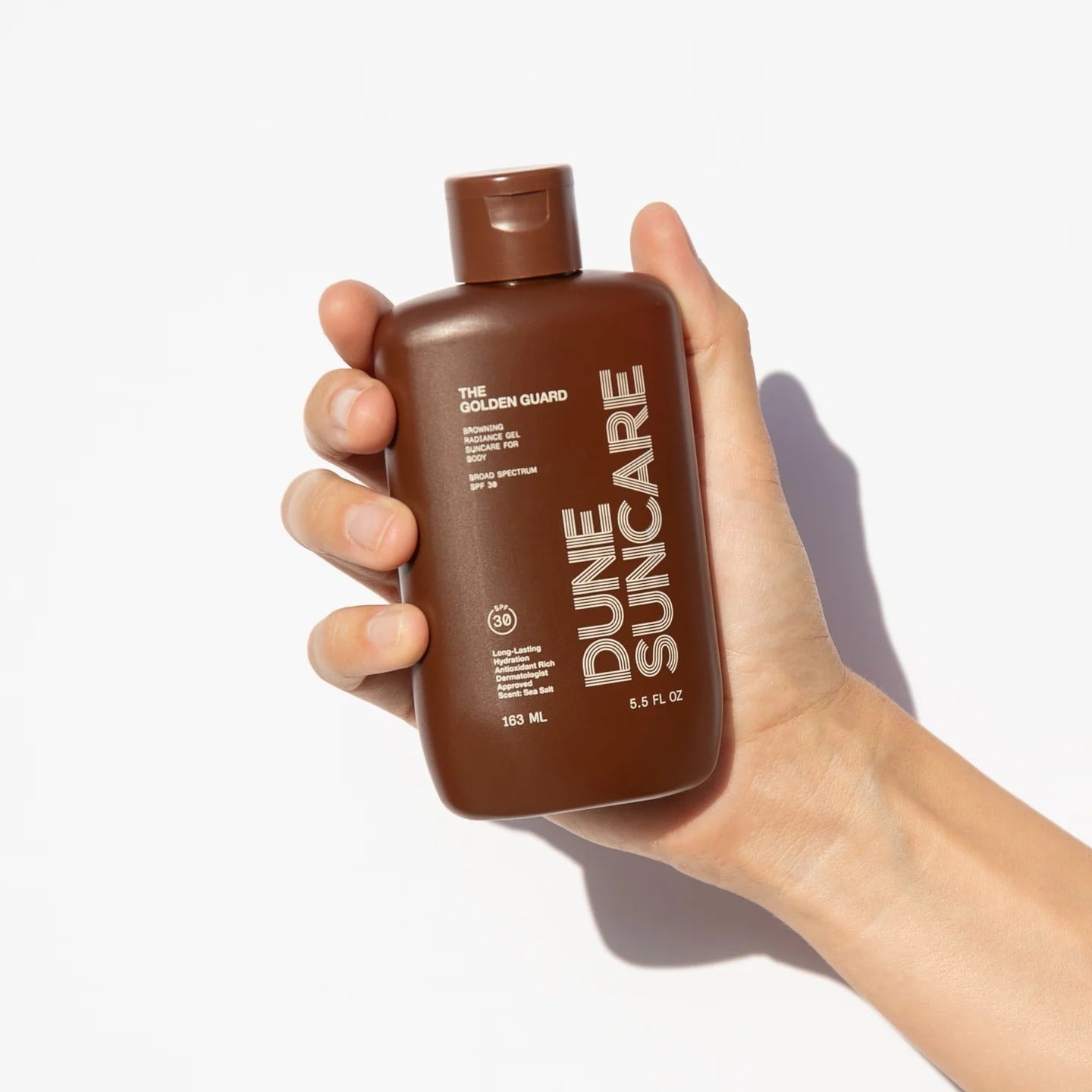 Hand holding a bottle of Dune Suncare sunscreen with a white background.
