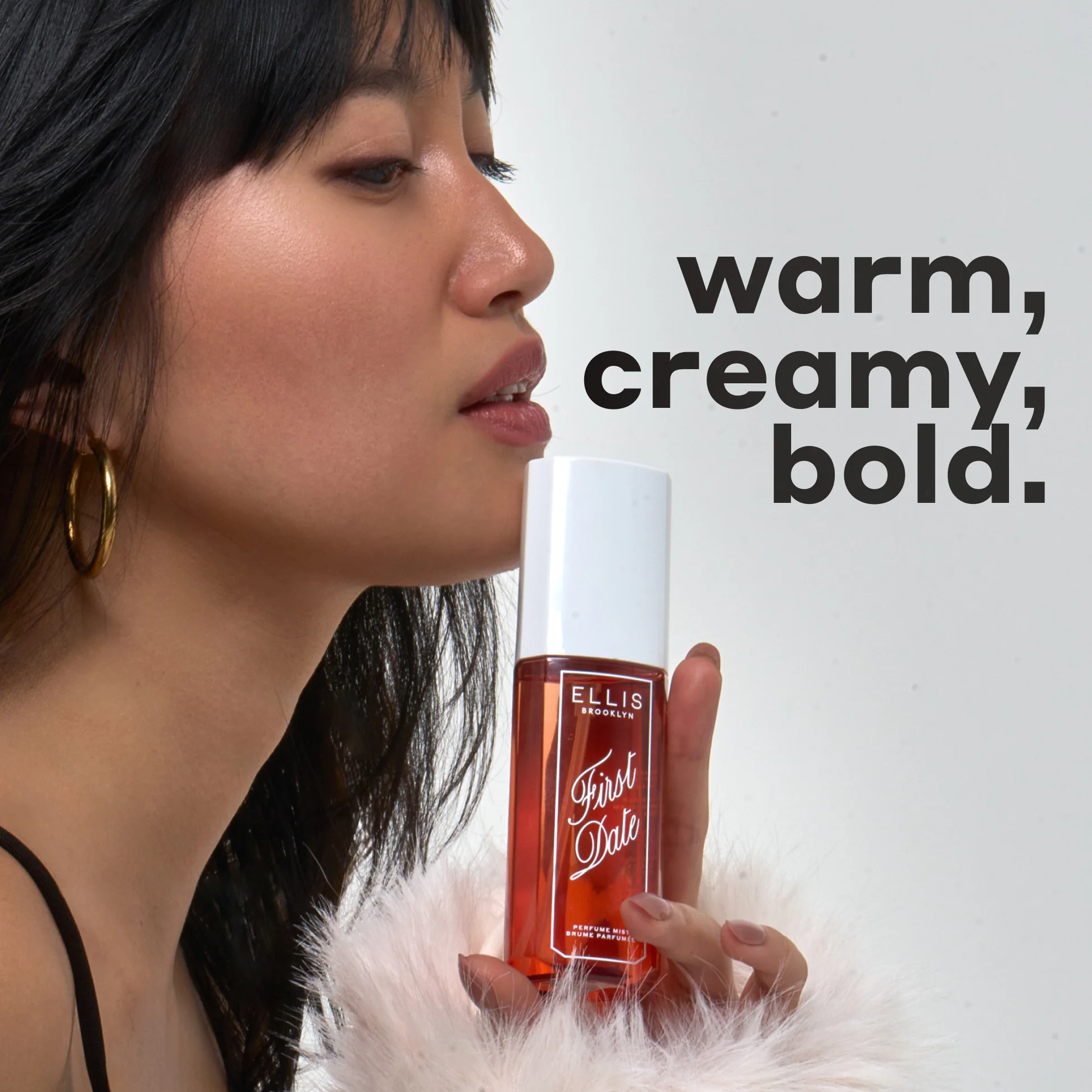 Woman holding a bottle of Ellis Brooklyn First Date perfume with text 'warm, creamy, bold' on a white background.
