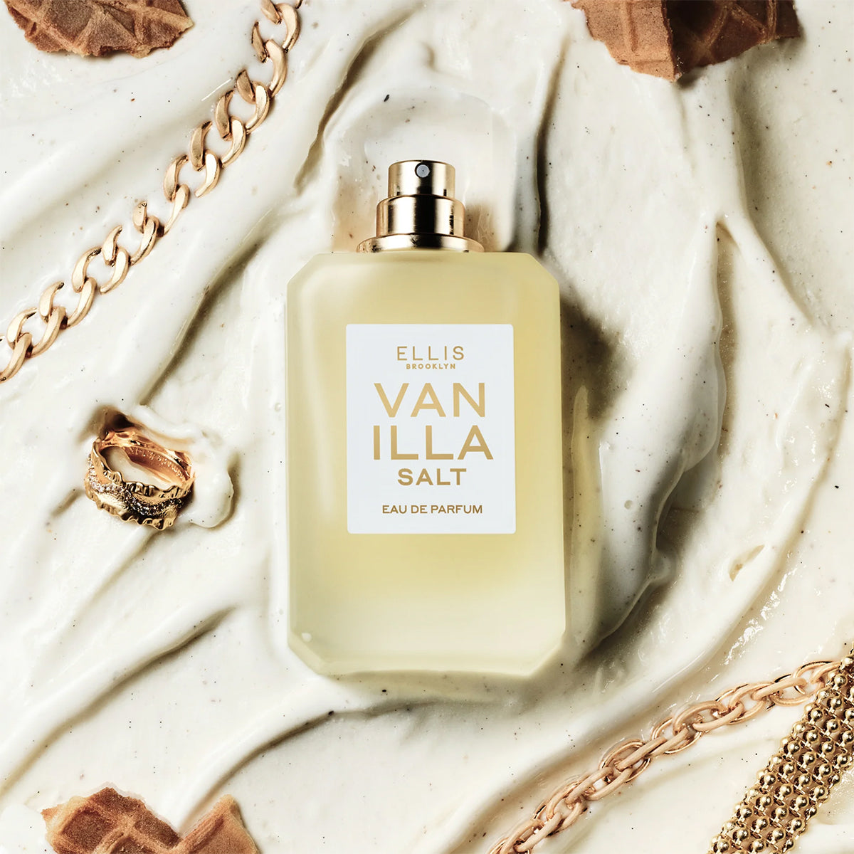 Perfume bottle labeled 'Vanilla Salt' by Ellis Brooklyn on a textured surface with gold chains and a ring.