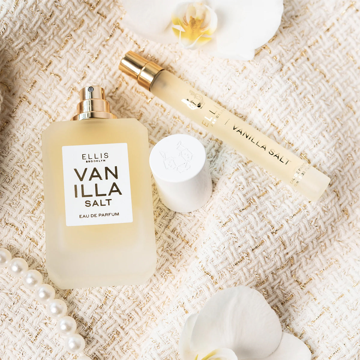 Perfume bottle labeled 'Vanilla Salt' by Ellis Brooklyn on a textured beige surface with flowers and pearls.