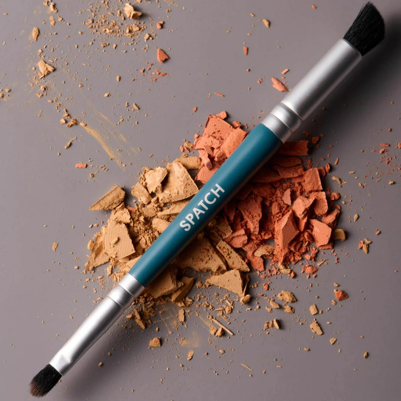 A makeup brush with a teal handle and silver ferrule, branded 'SPATCH', laid diagonally across crushed pressed powder in tan and terracotta shades on a neutral gray background.